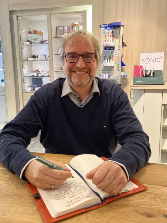 Jens König sitzt  mit einem Notizbuch an einem Holztisch im SchreibZimmer vor den Schreibgerätevitrinen